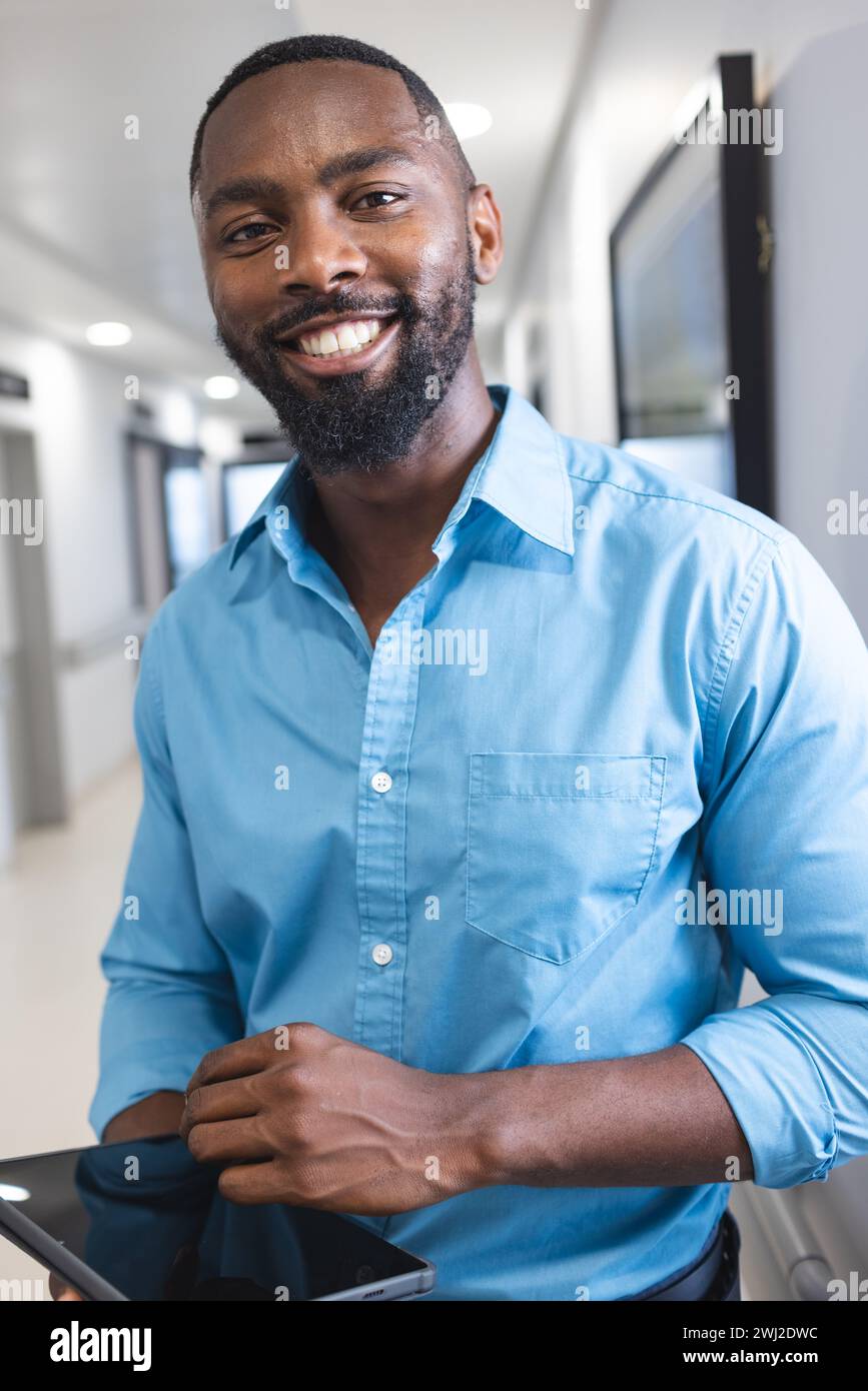 Retrato del médico masculino afroamericano feliz que sostiene la tableta en el pasillo en el hospital Foto de stock