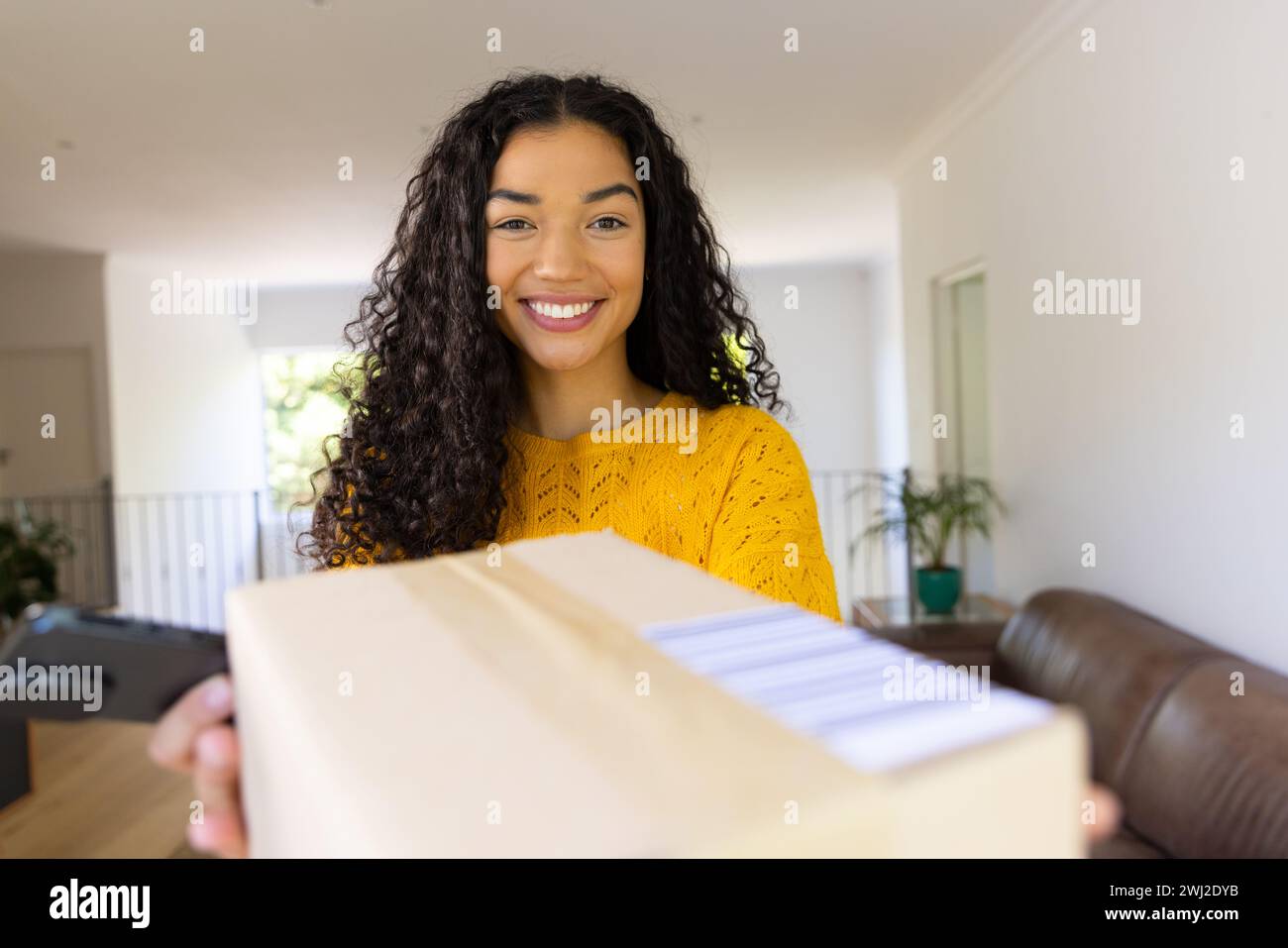 Mujer biracial feliz en suéter amarillo que sostiene el paquete y que usa el teléfono inteligente en casa Foto de stock