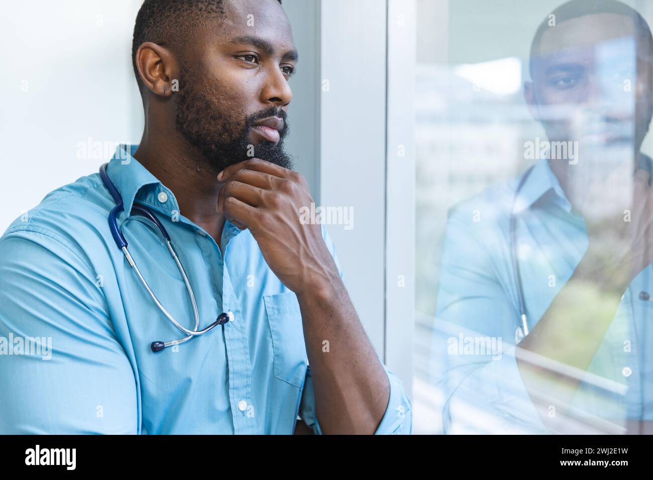 Pensativo médico masculino afroamericano con camisa azul mirando a través de la ventana en la habitación del hospital Foto de stock
