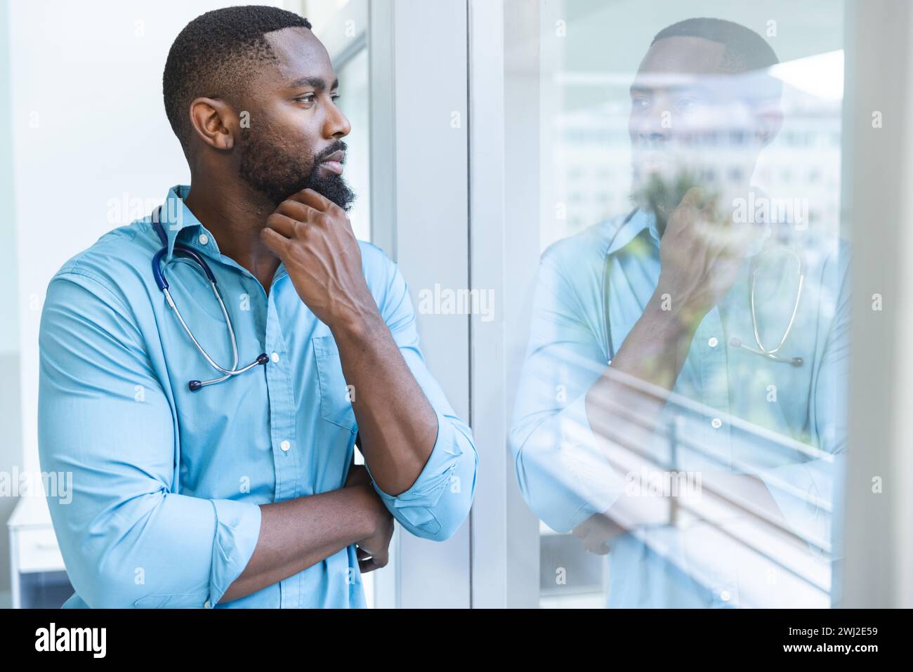 Pensativo médico masculino afroamericano con camisa azul mirando a través de la ventana en la habitación del hospital Foto de stock