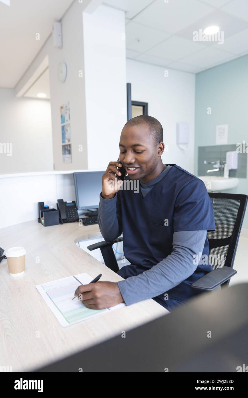 Médico masculino afroamericano hablando en teléfono inteligente y tomando notas en el mostrador de recepción en el hospital Foto de stock