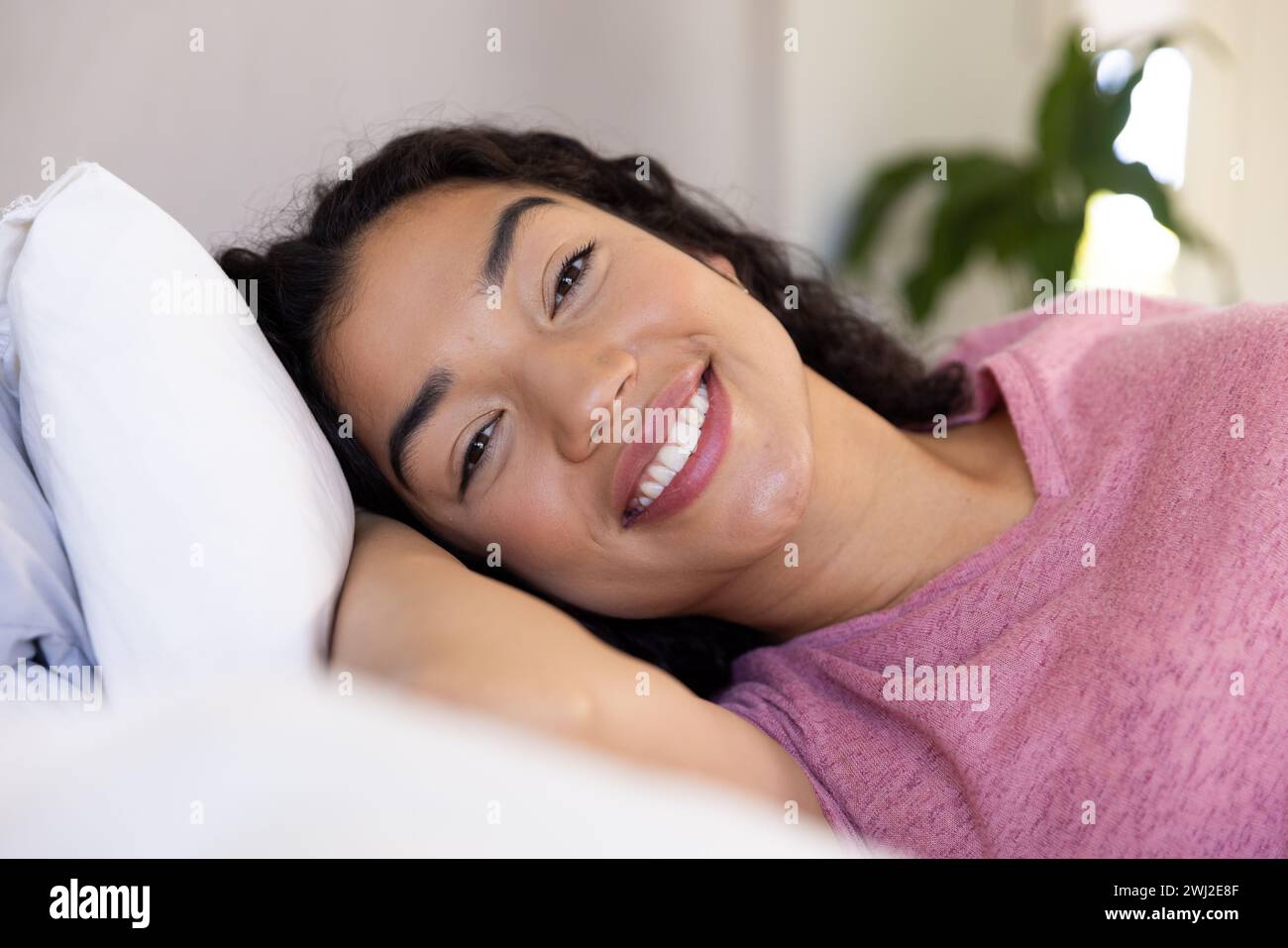 Mujer biracial feliz acostada en la cama en casa soleada Foto de stock