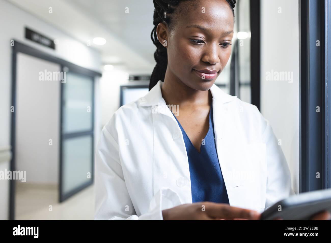 Médico afroamericano con bata de laboratorio usando tableta en el pasillo del hospital Foto de stock