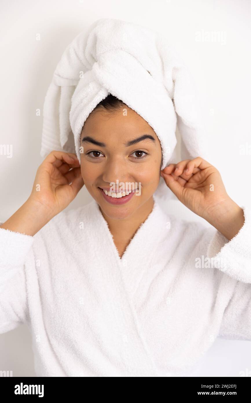 Retrato de mujer biracial feliz en albornoz y con toalla contra la pared blanca Foto de stock