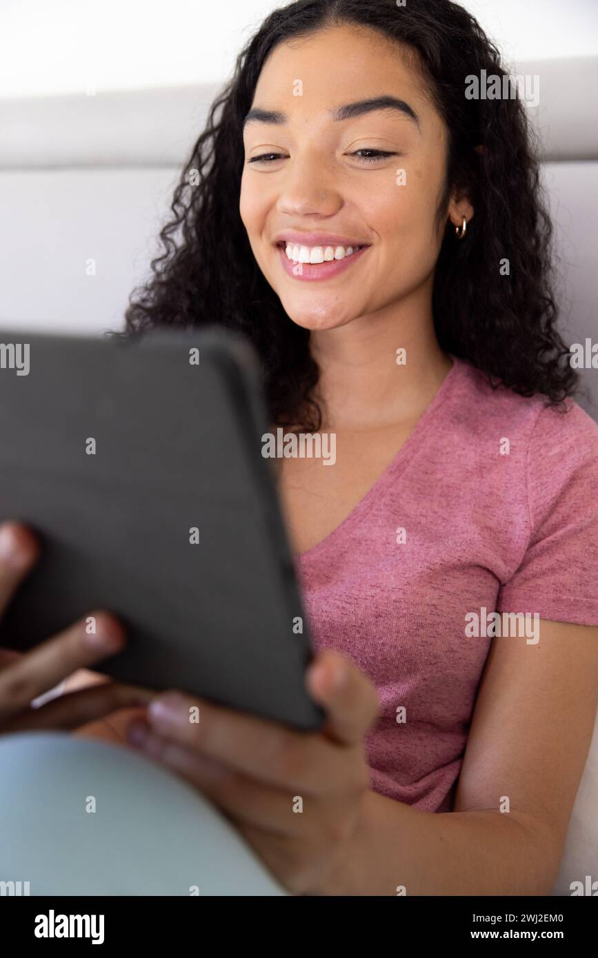 Mujer biracial feliz usando tableta sentada en la cama en casa soleada Foto de stock