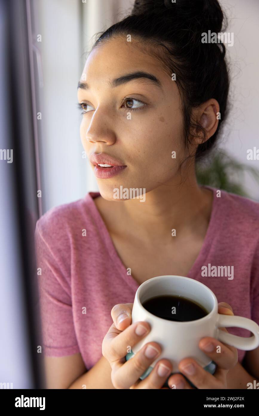 Mujer biracial sosteniendo taza de café y mirando por la ventana en casa soleada Foto de stock