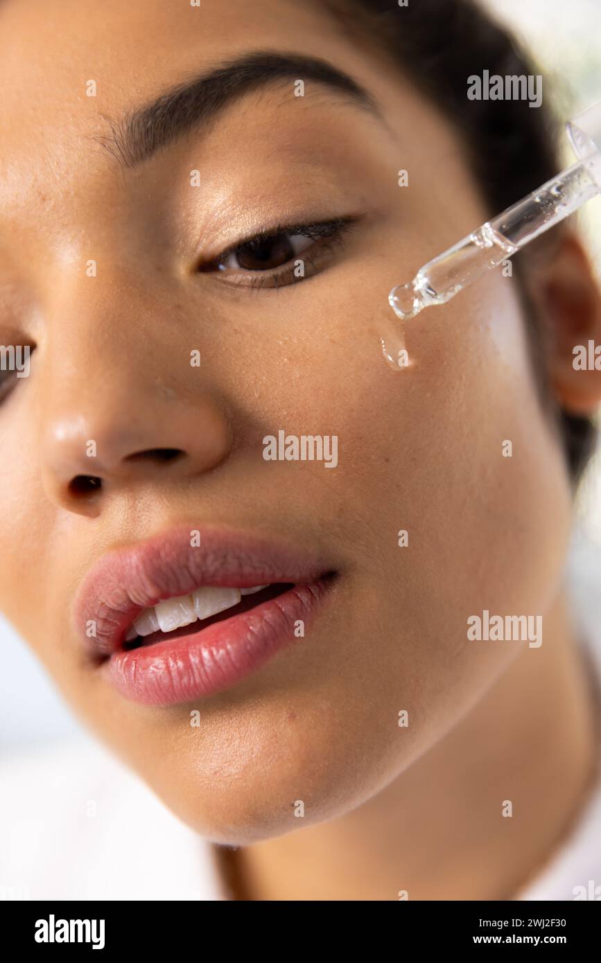 Mujer biracial en albornoz aplicando cosmética en su cara en baño soleado Foto de stock