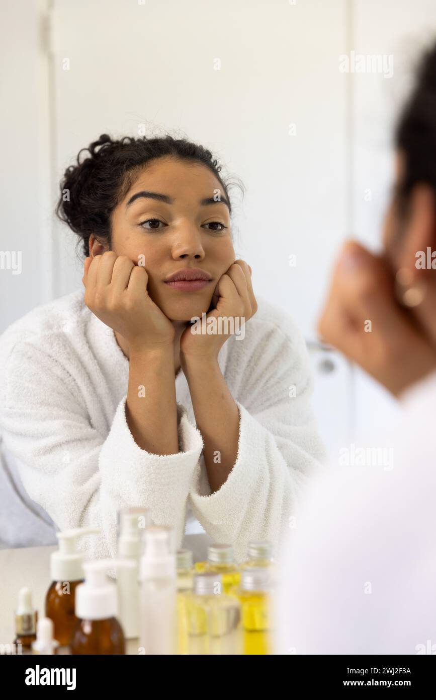 Mujer biracial en albornoz mirando en espejo en baño soleado Foto de stock