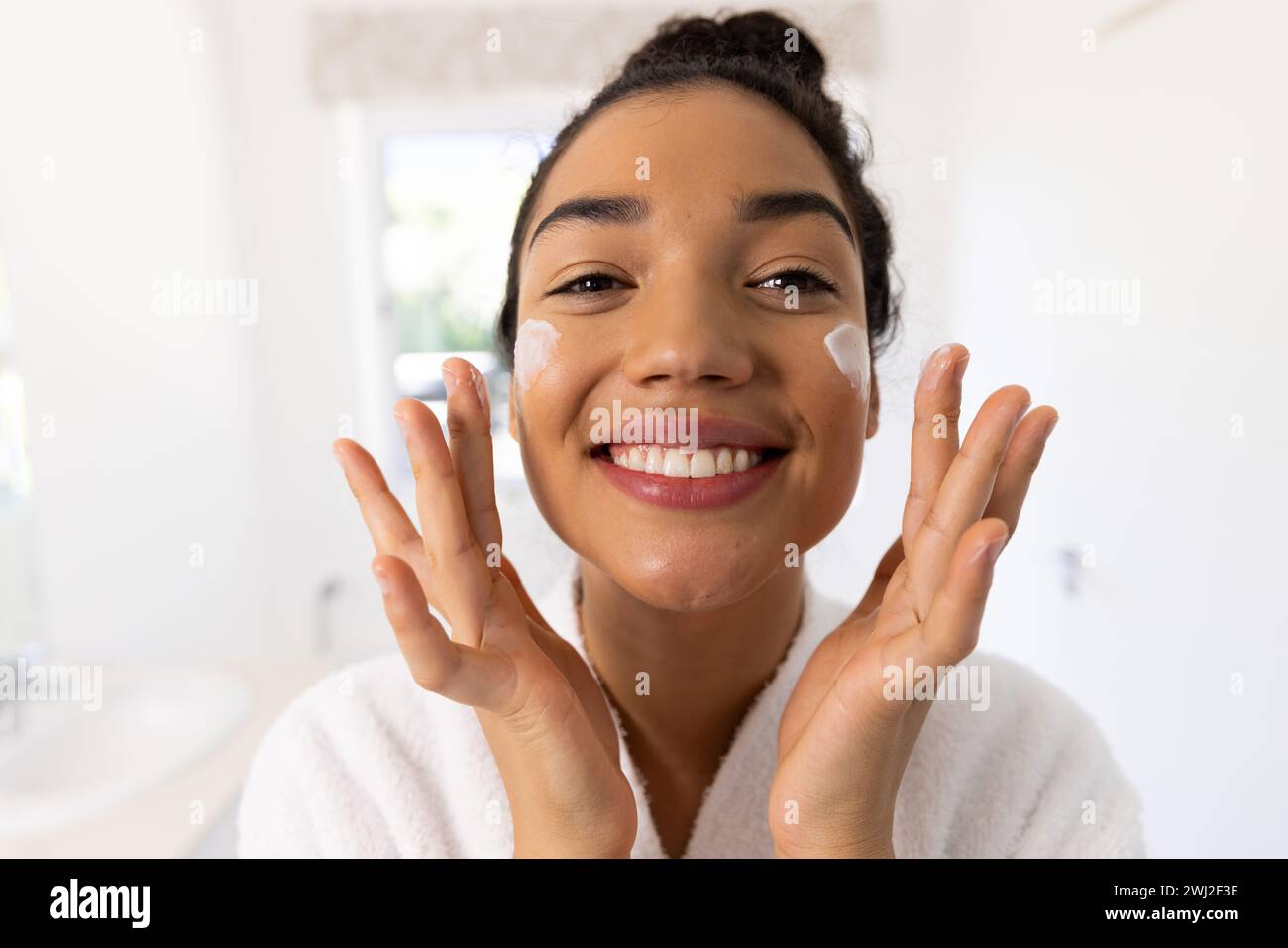 Retrato de mujer biracial feliz en albornoz aplicando crema en su cara en baño soleado Foto de stock