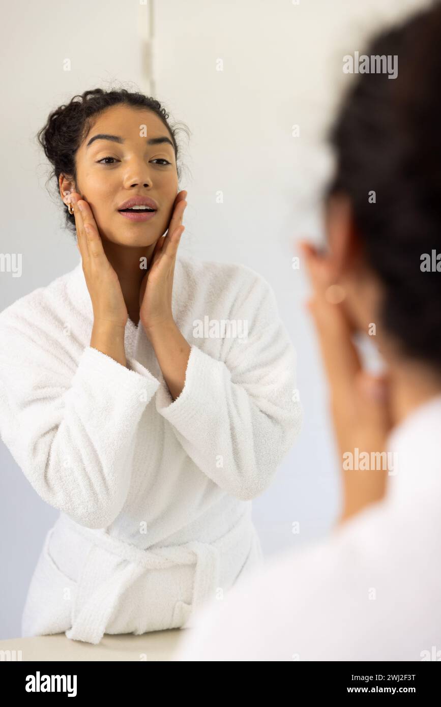 Mujer biracial en albornoz mirando en espejo en baño soleado Foto de stock