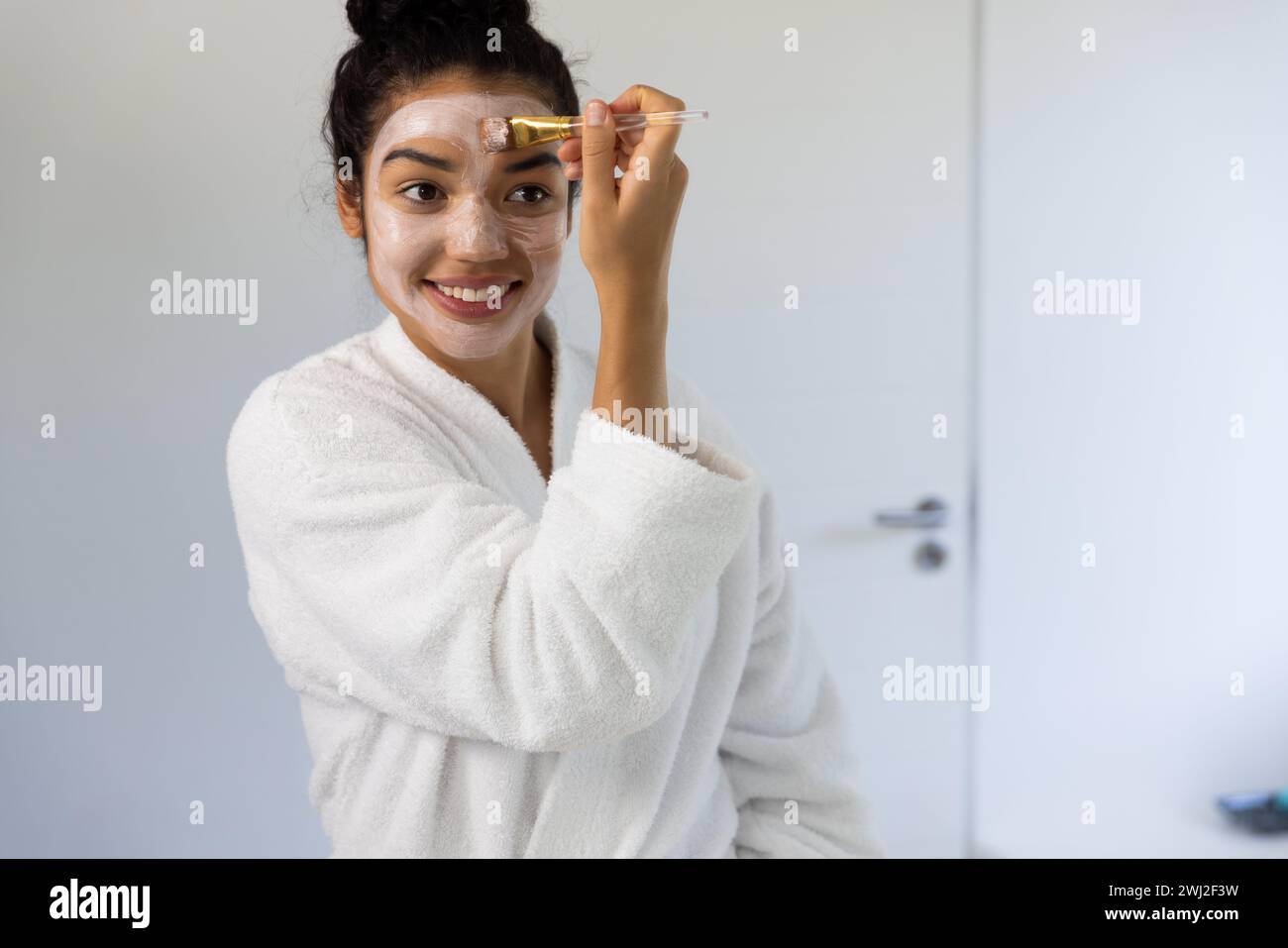 Mujer biracial feliz en albornoz aplicando máscara facial usando cepillo en baño soleado Foto de stock
