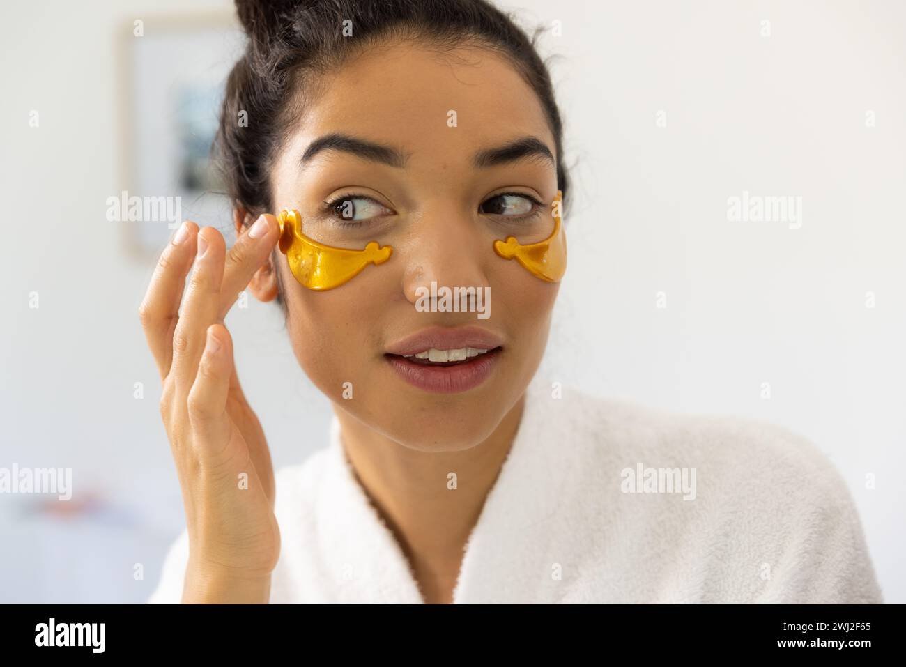 Mujer biracial en albornoz aplicando máscara de ojos en baño soleado Foto de stock