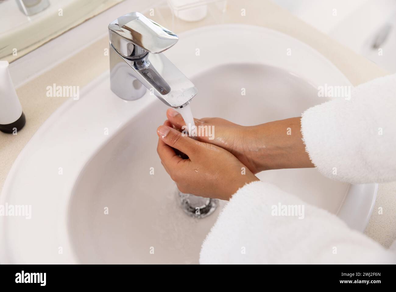 Lavarse las manos de la mujer biracial en albornoz en baño soleado Foto de stock