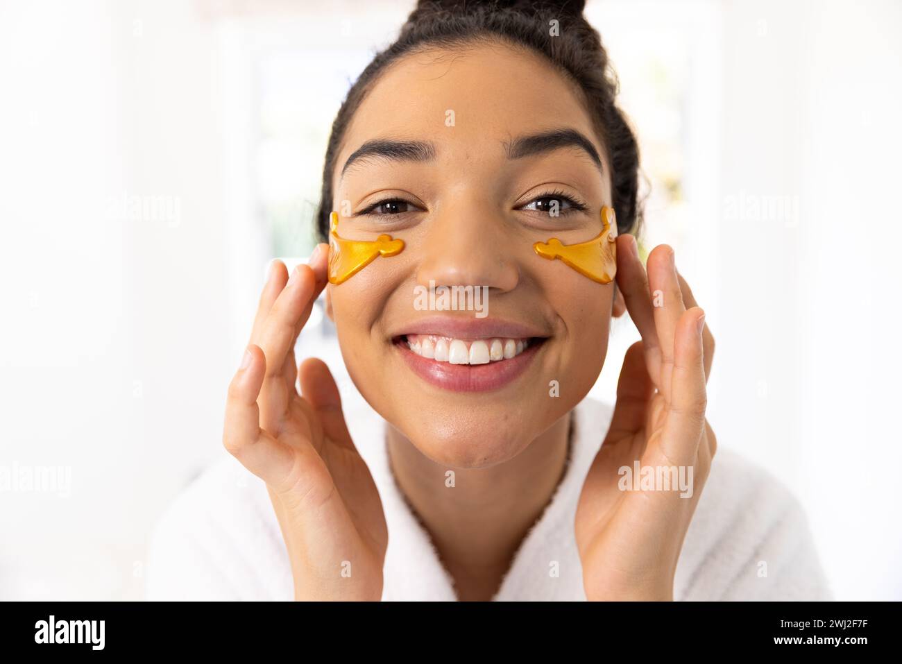 Retrato de mujer biracial feliz en albornoz aplicando máscara de ojo en baño soleado Foto de stock