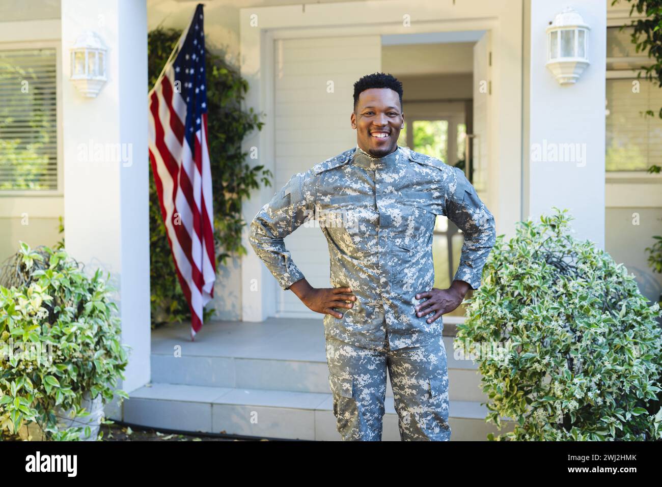 Soldado masculino afroamericano feliz con las manos en las caderas fuera de casa con la bandera de ee.uu., espacio de copia Foto de stock