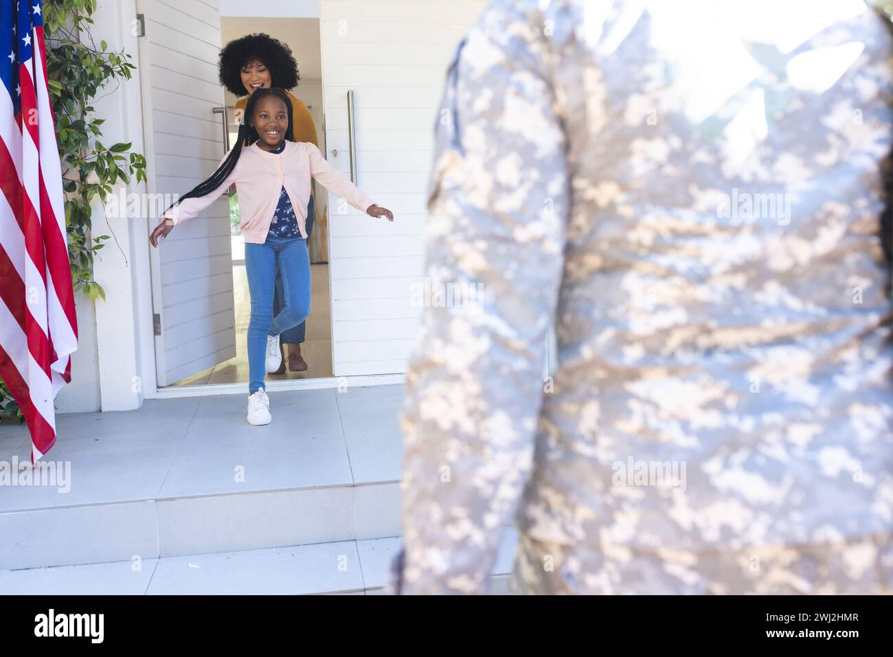 Feliz esposa e hija afroamericana dando la bienvenida al soldado masculino al aire libre con la bandera de ee.uu., espacio de copia Foto de stock