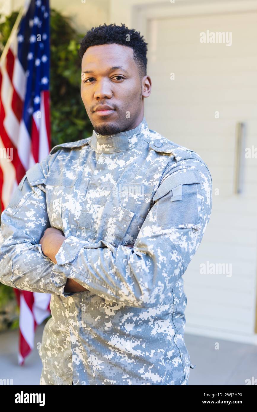 Soldado masculino afroamericano de pie con los brazos cruzados fuera de casa con la bandera de ee.uu., espacio de copia Foto de stock