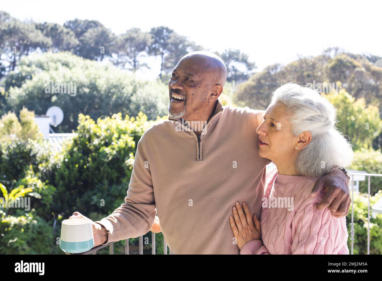 Feliz pareja senior diversa sosteniendo una taza de café y abrazando en la terraza soleada Foto de stock