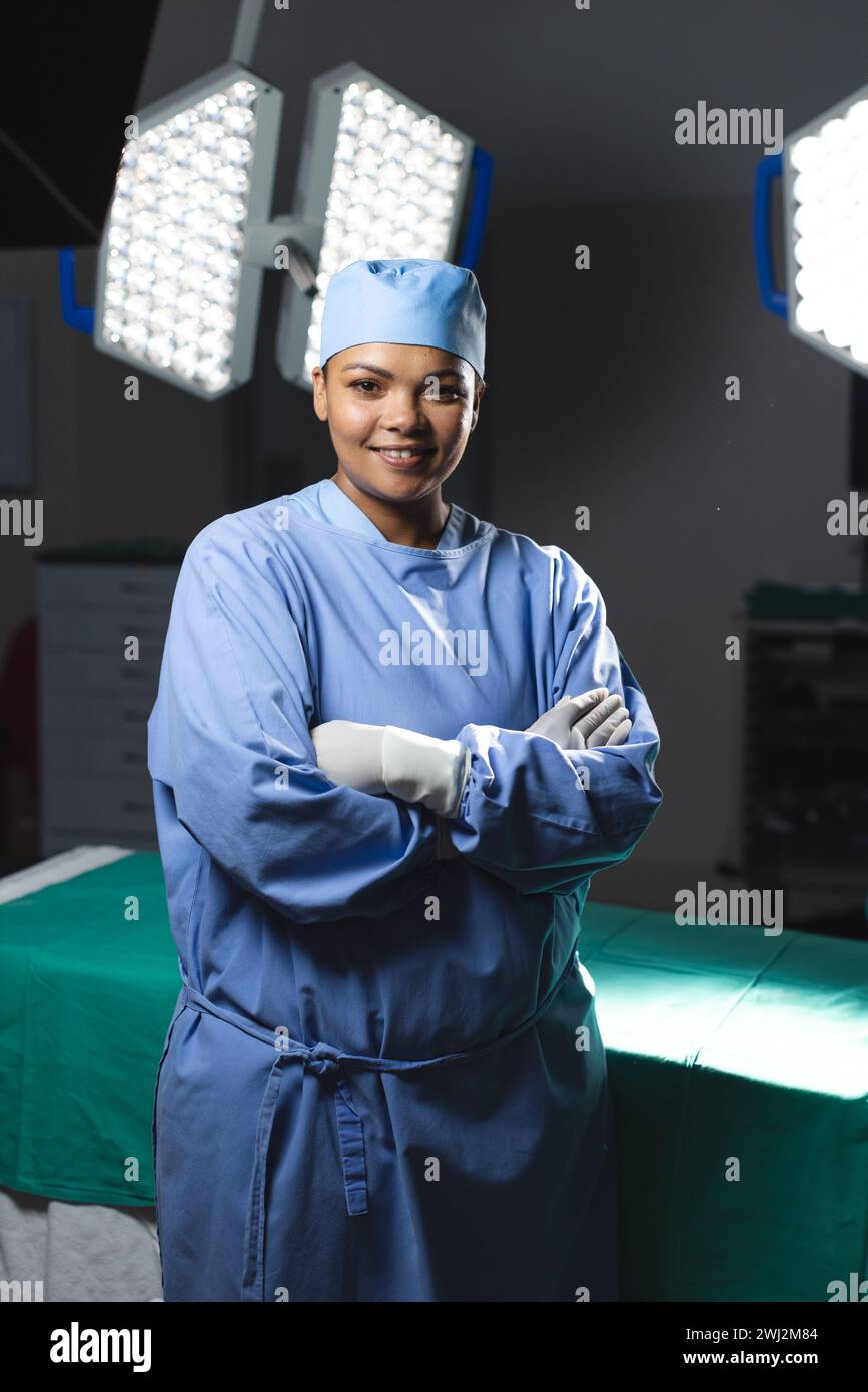 Retrato de cirujano femenino biracial feliz que lleva bata quirúrgica en el quirófano Foto de stock