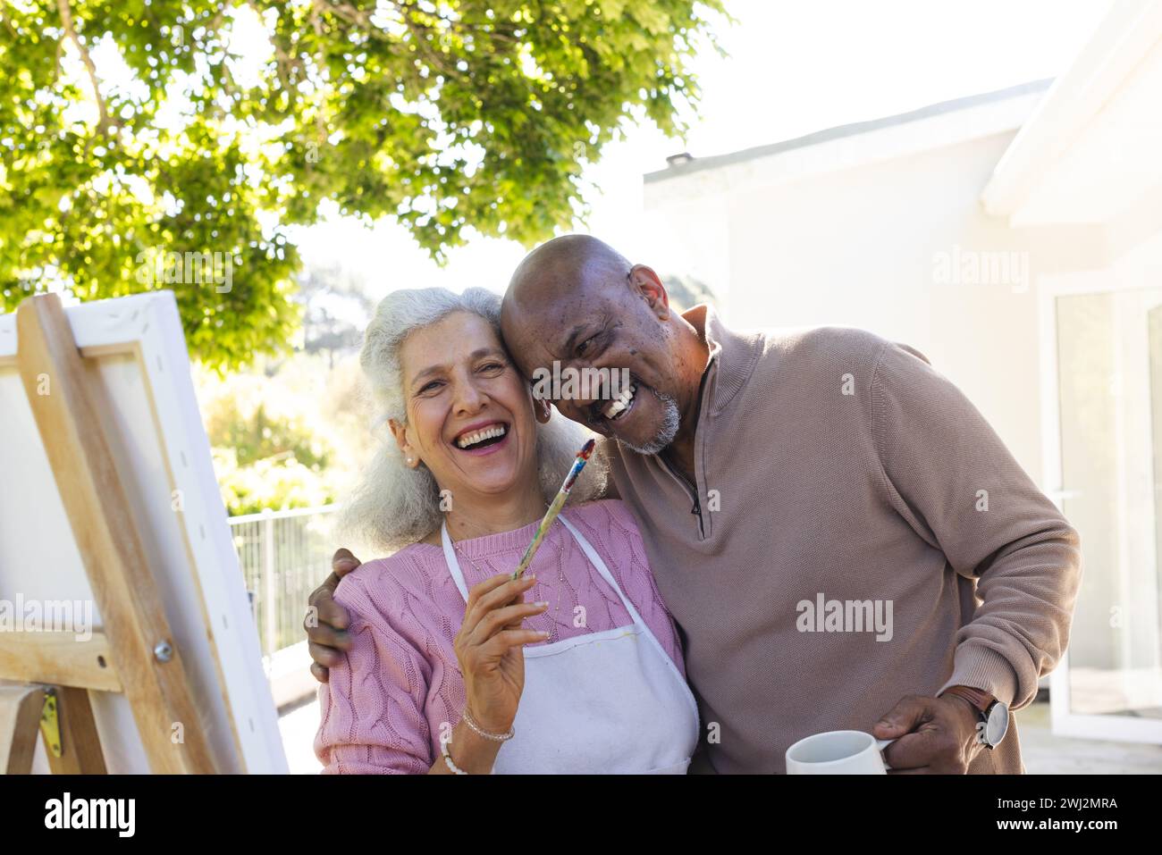Feliz pareja senior diversa abrazando y mirando la pintura en la terraza soleada Foto de stock