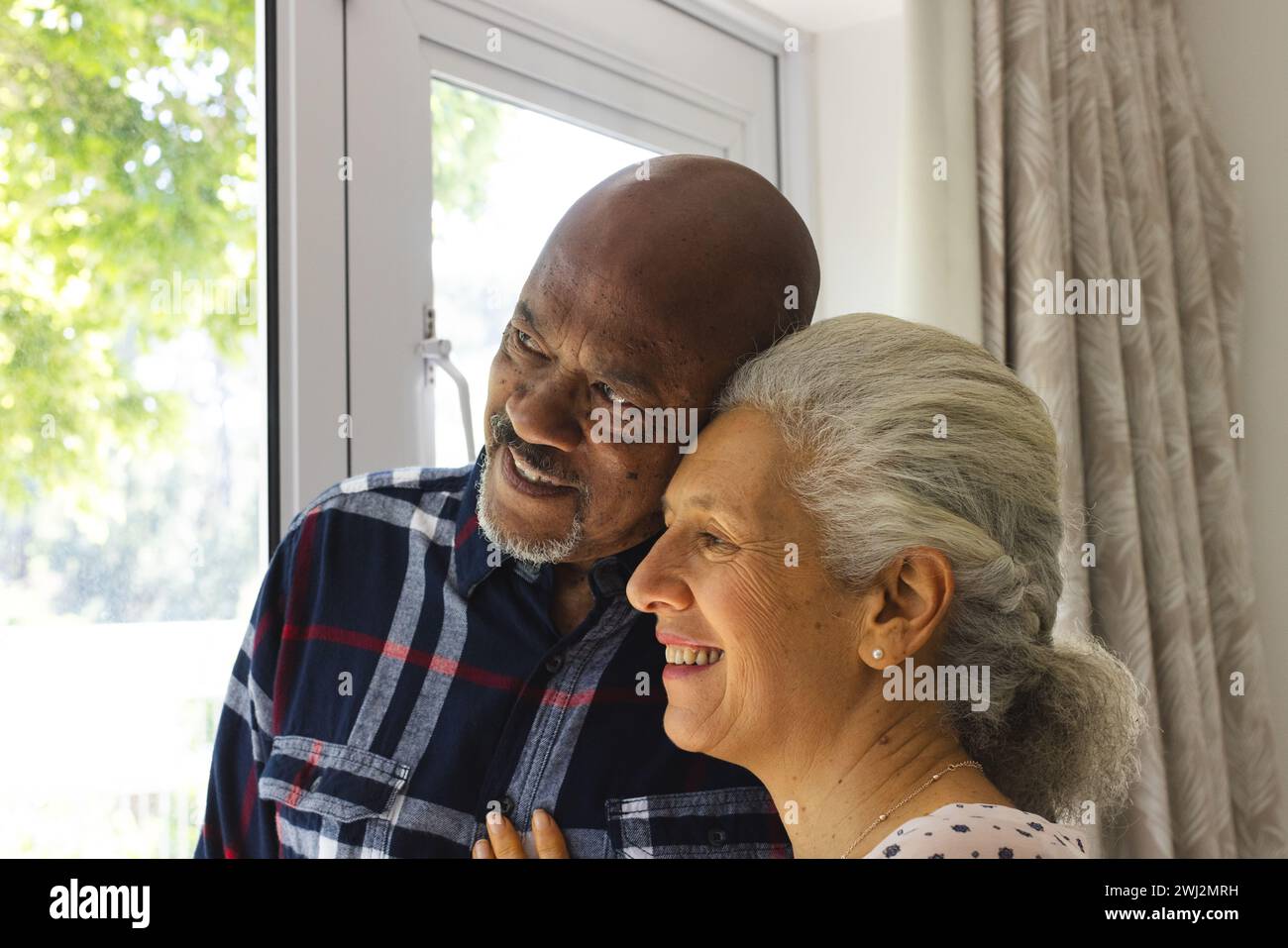 Feliz pareja senior diversa abrazando y mirando por la ventana en casa soleada Foto de stock