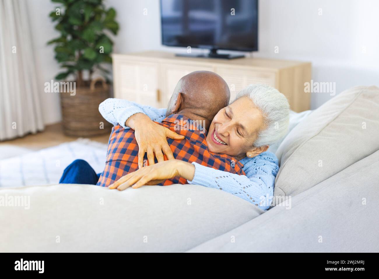 Feliz pareja senior diversa sentada en el sofá y abrazando en la sala de estar soleada Foto de stock