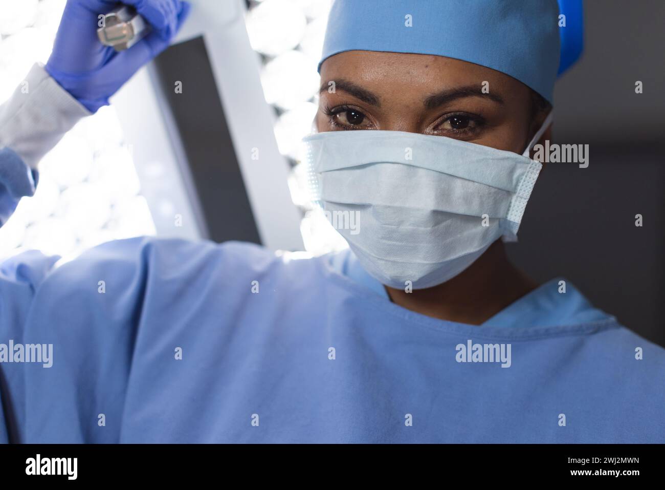 Retrato de cirujano femenino biracial con bata quirúrgica y máscara facial en quirófano Foto de stock