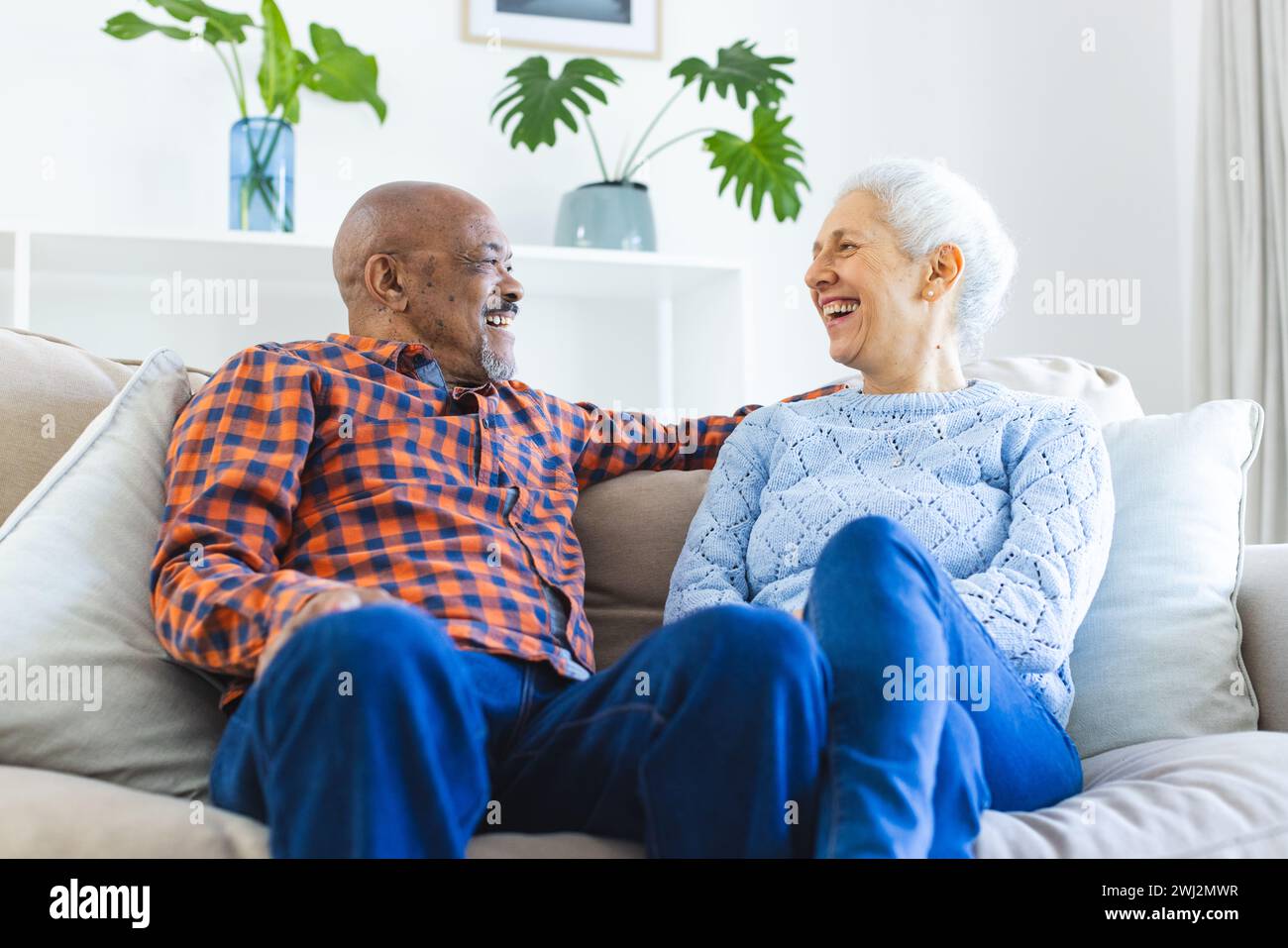 Feliz pareja senior diversa sentada en el sofá, hablando y abrazando en la sala de estar soleada Foto de stock