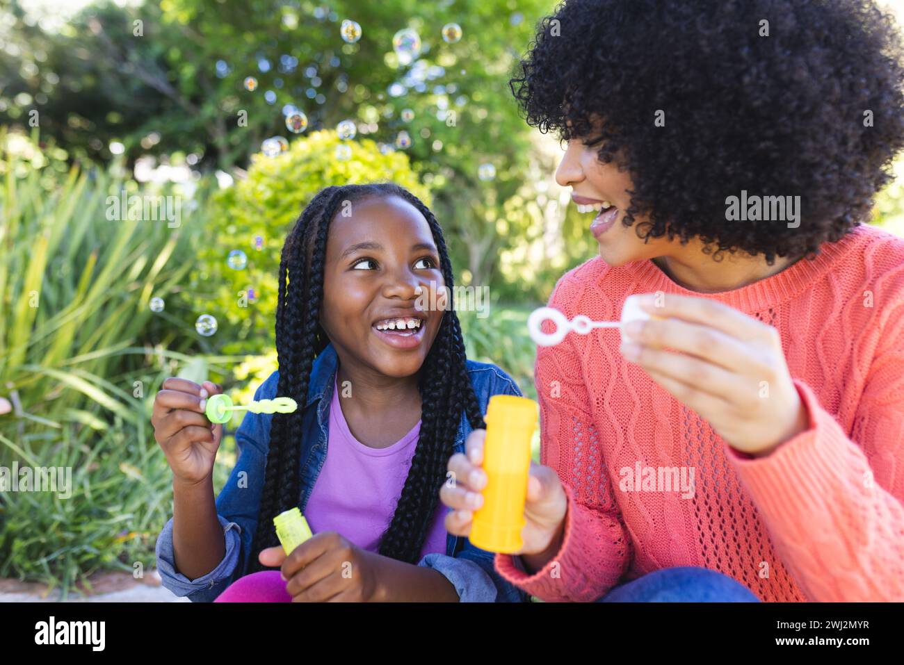 Feliz madre e hija afroamericana sentada y soplando burbujas en jardín soleado, espacio de copia Foto de stock