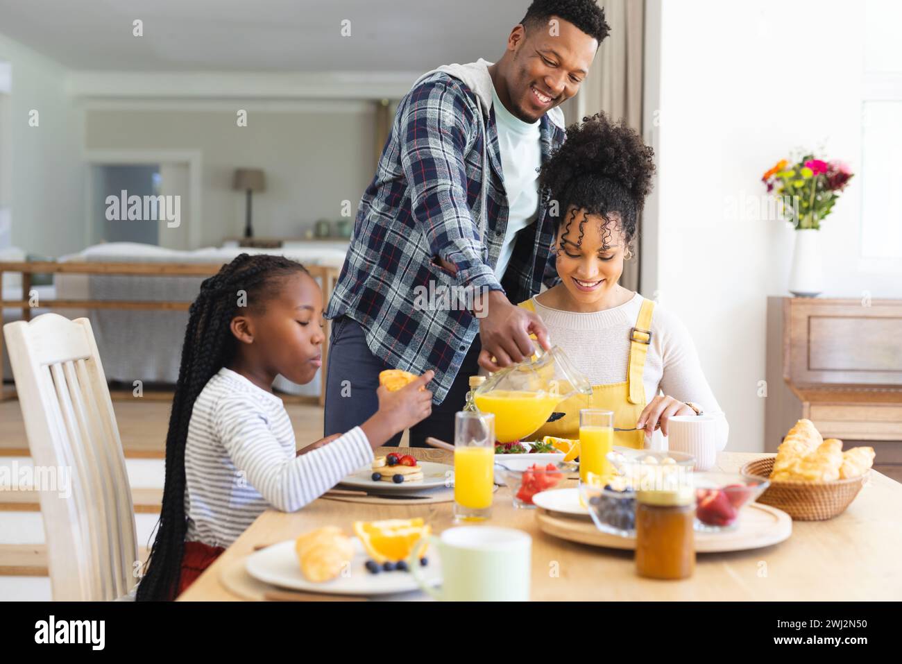 Hombre afroamericano feliz que vierte jugo de naranja en el vidrio en la mesa en casa, espacio de copia Foto de stock