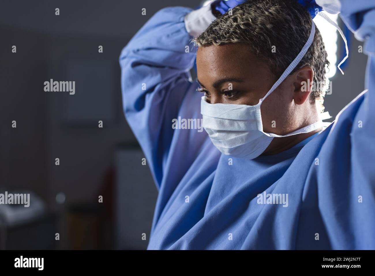 Cirujano femenino biracial que lleva bata quirúrgica y máscara facial en quirófano Foto de stock