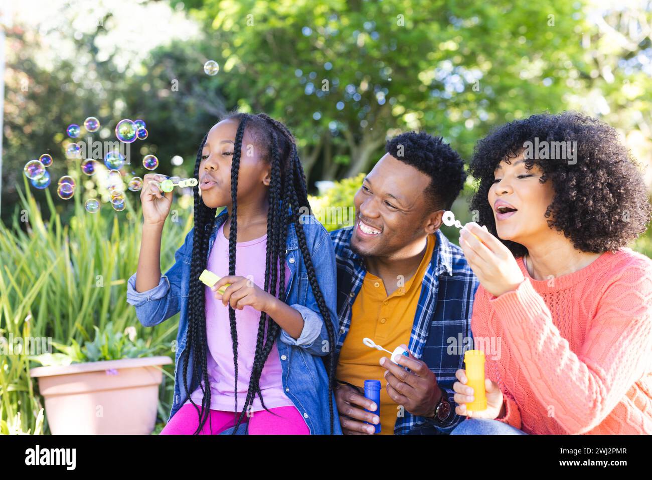 Padre afroamericano feliz, madre e hija que sopla burbujas en jardín soleado, espacio de copia Foto de stock