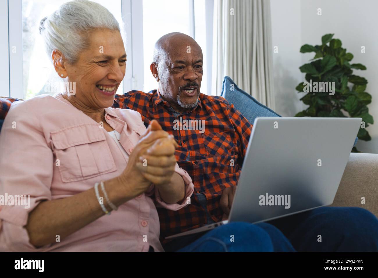 Feliz pareja senior diversa que se sienta en el sofá, abrazando y usando el ordenador portátil en la sala de estar soleada Foto de stock