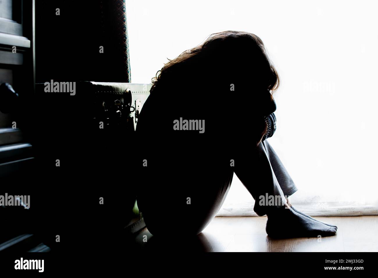 Infeliz mujer madura, mujer en silueta, sentado en el suelo con la espalda iluminada. Pensamientos deprimentes. Foto de stock