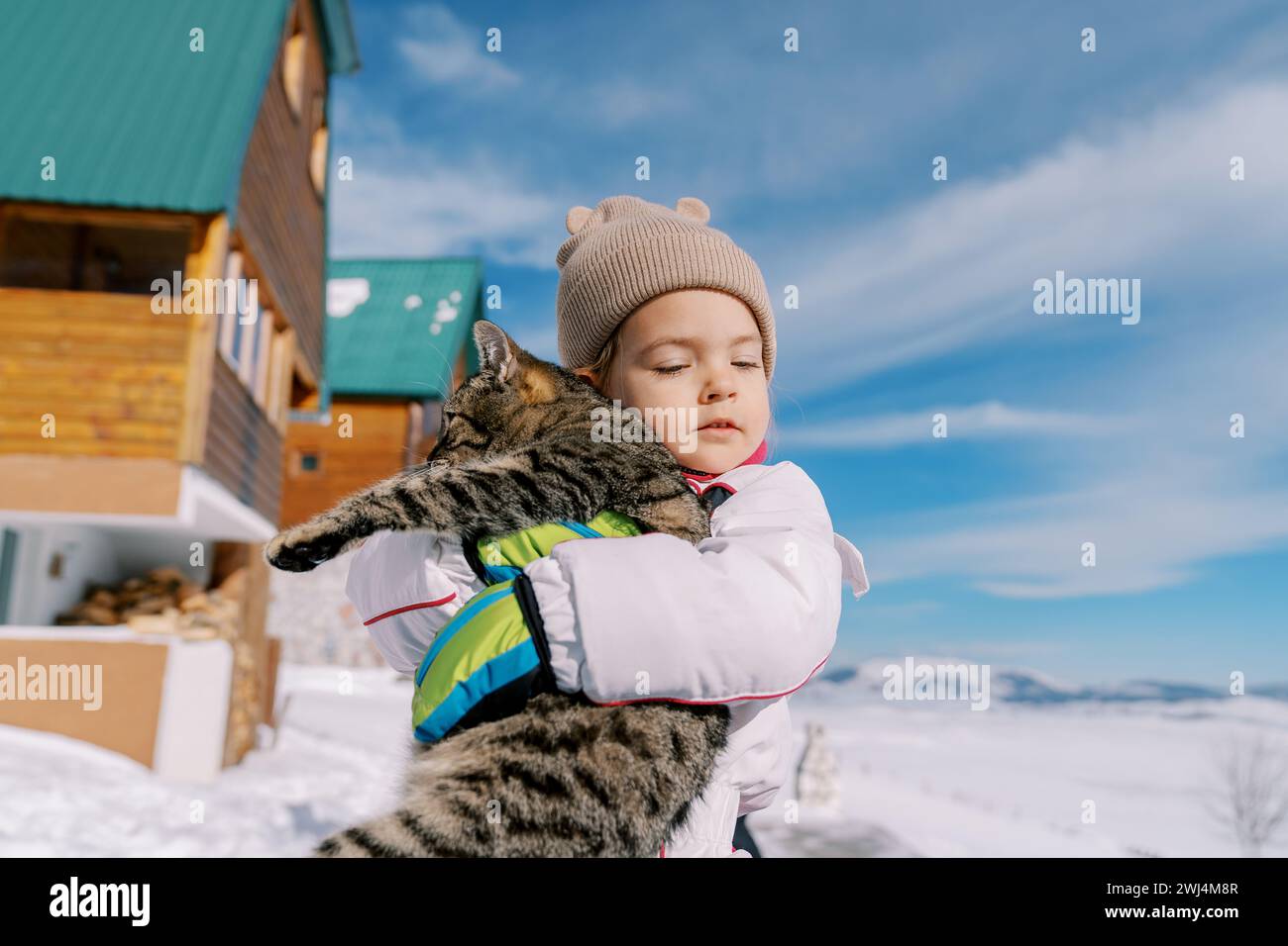 La niña abraza a un gato tabby mientras está de pie en la nieve cerca de una cabaña de madera Foto de stock