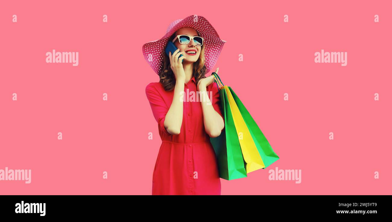 Hermosa mujer joven feliz que llama en el teléfono móvil con el bolso de compras que mira lejos en sombrero de verano en fondo de estudio rosa Foto de stock