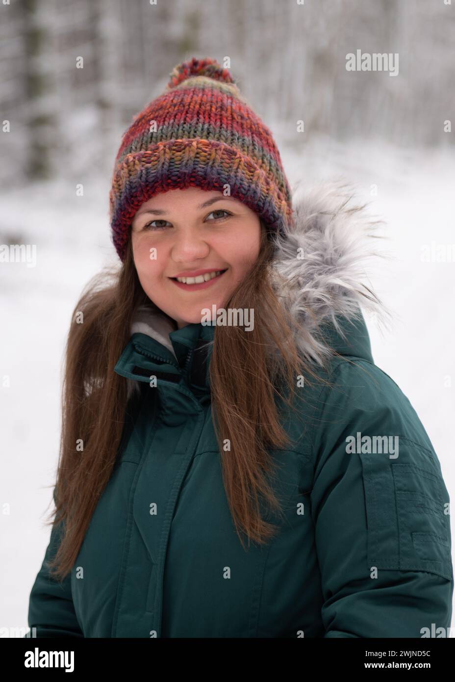 una mujer de cara amistosa con un sombrero de invierno vibrante y una chaqueta verde azulada, contra el telón de fondo sereno de un bosque nevado. Su cálida sonrisa contrasta con t Foto de stock