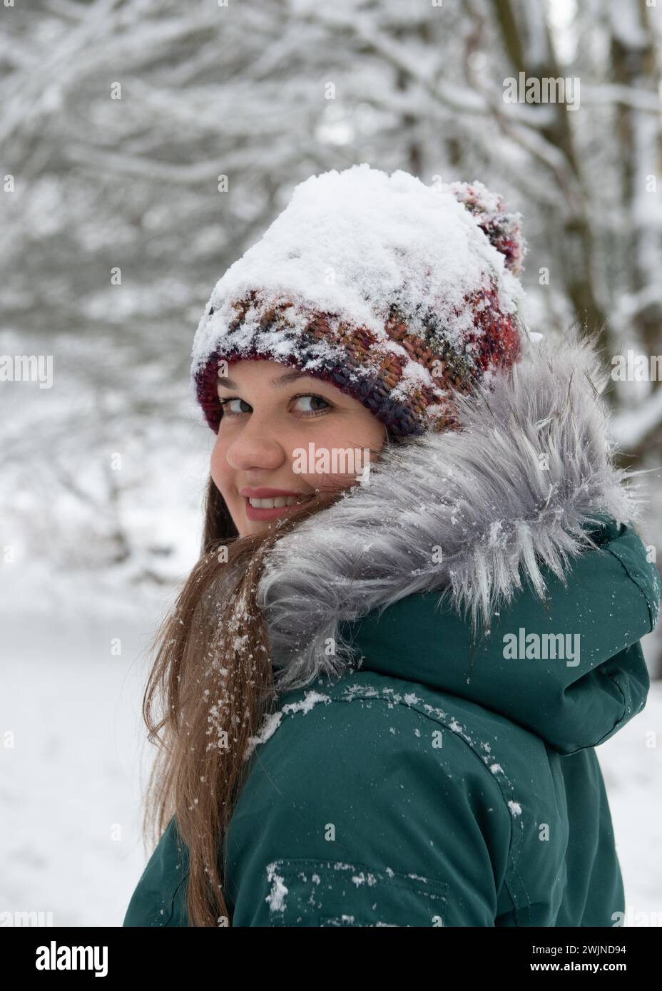 una mujer que ofrece una sonrisa cálida, girando hacia la cámara con su sombrero cubierto de una capa fresca de nieve. Ella está vestida para el frío en un invierno verde Foto de stock