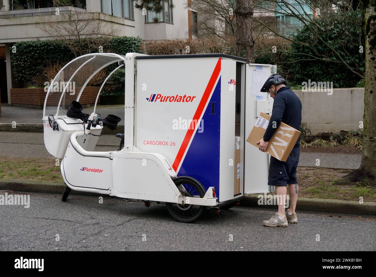 Repartidor de pie junto a su bicicleta de carga eléctrica Purolator estacionada en una calle residencial en Vancouver, BC, Canadá Foto de stock