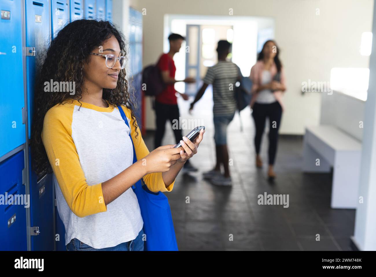Joven mujer biracial revisa su teléfono en un pasillo de la escuela secundaria con espacio de copia Foto de stock