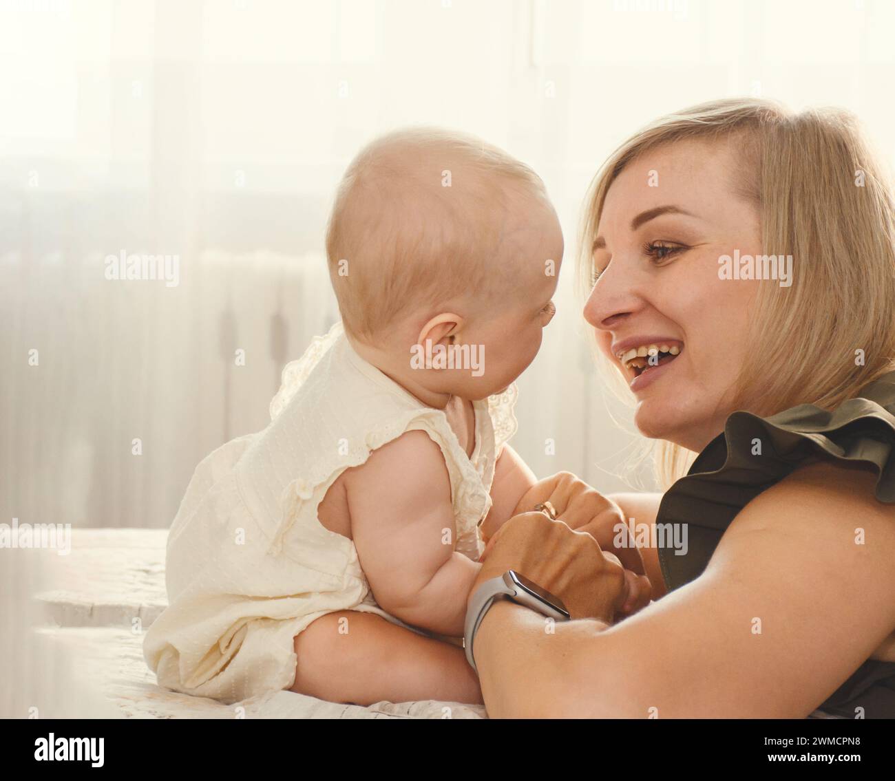 joven madre juega, coos y ríe con su bebé en un día soleado en casa Foto de stock