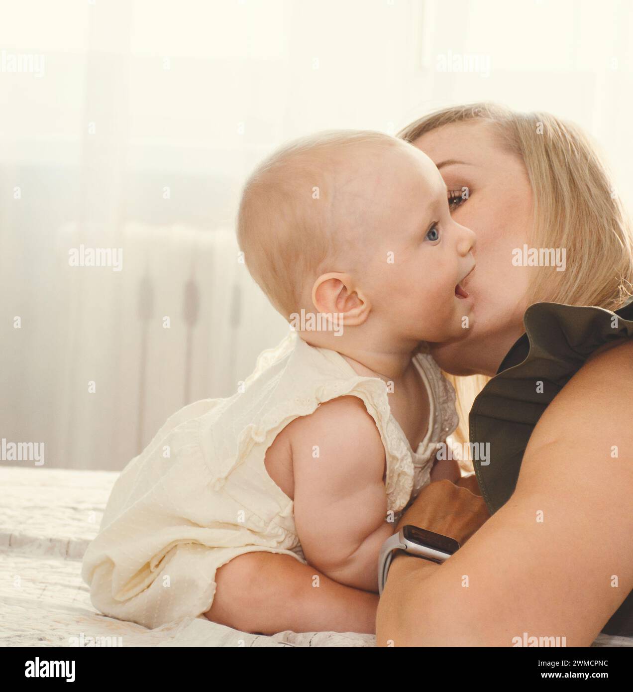 la joven madre besa a su bebé en la mejilla en un día soleado en casa Foto de stock