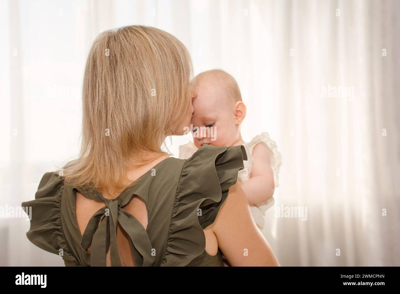 la joven madre sostiene a su bebé en sus brazos y la besa, en un día soleado en casa, enfoque suave en la mamá Foto de stock
