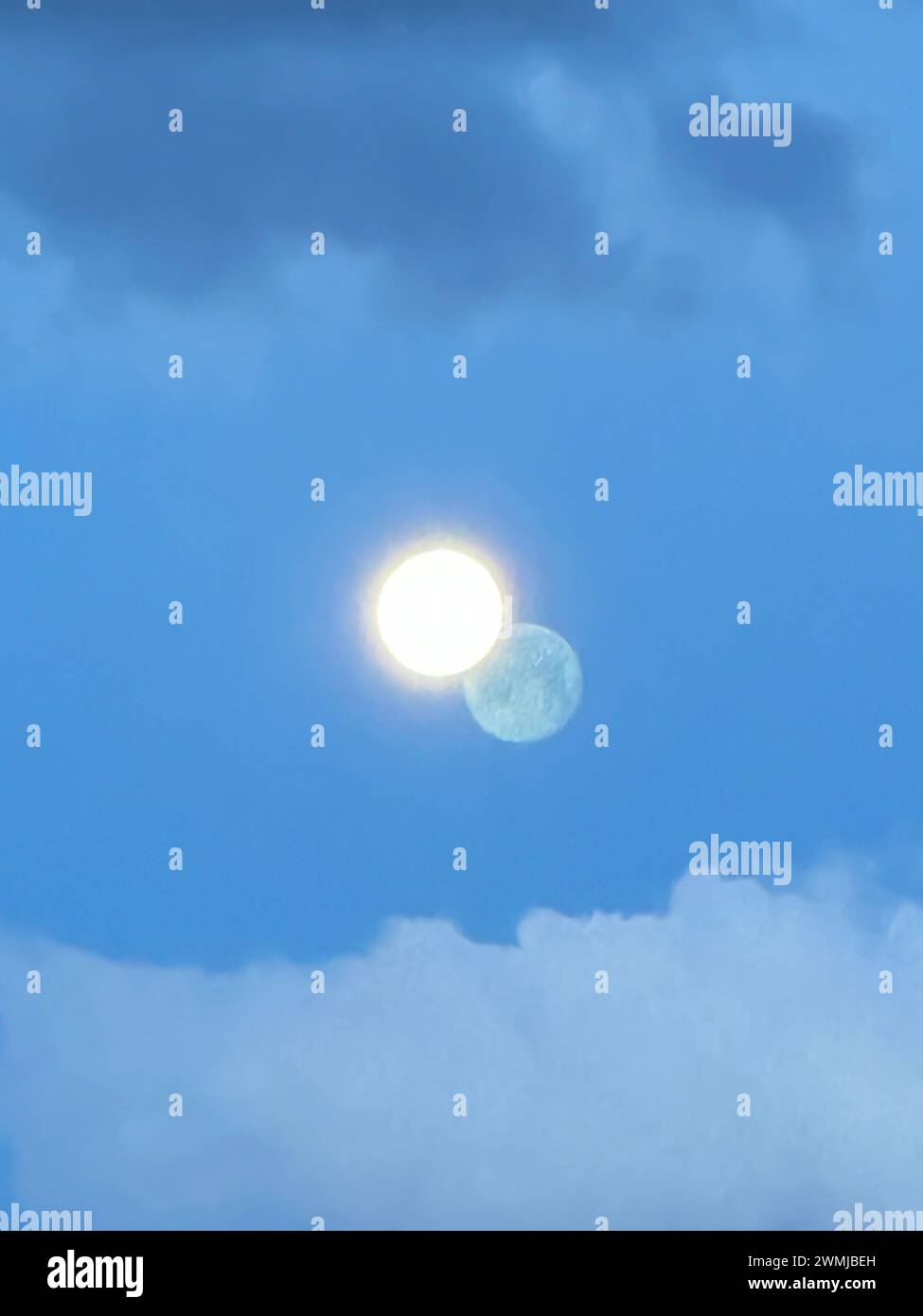 Una luna brillante que brilla contra un cielo azul con una imagen de sombra en las nubes distantes parece una luna doble con un círculo amarillo o dos planetas distantes. Foto de stock