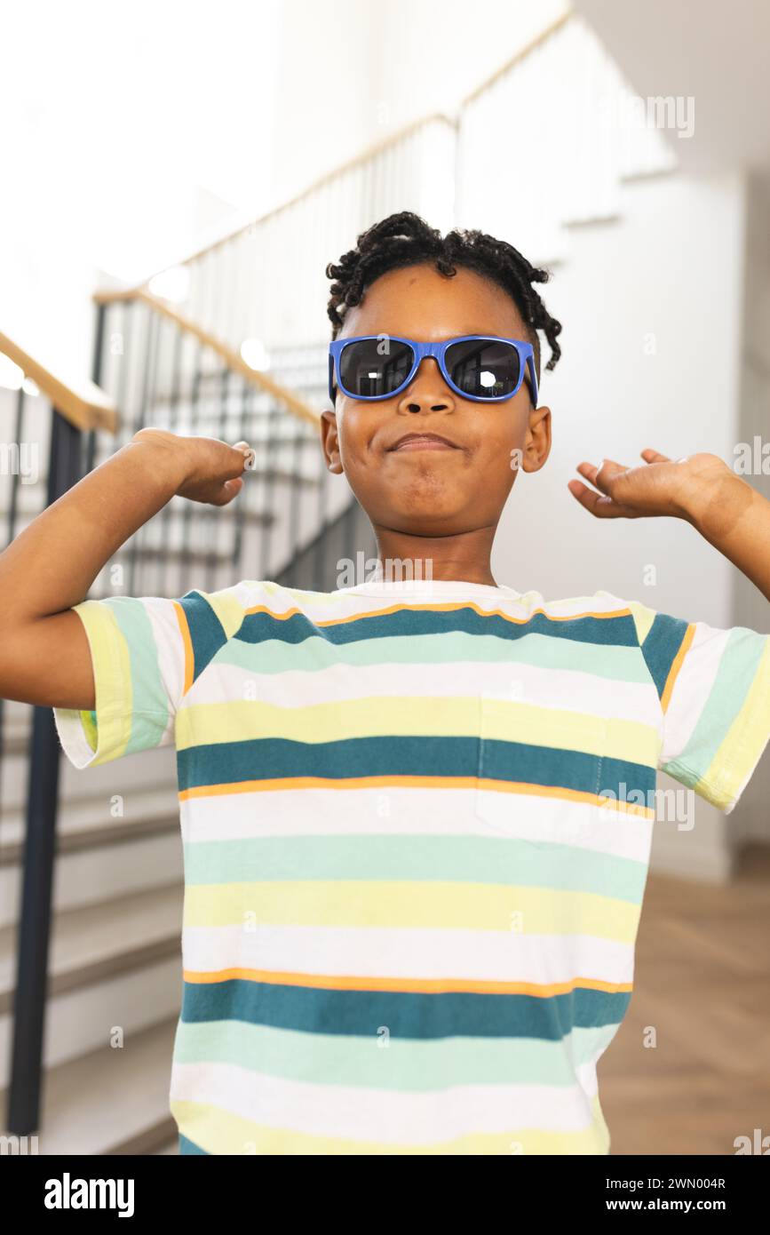 Niño afroamericano flexiona sus músculos, usando gafas de sol y una camisa de rayas Foto de stock