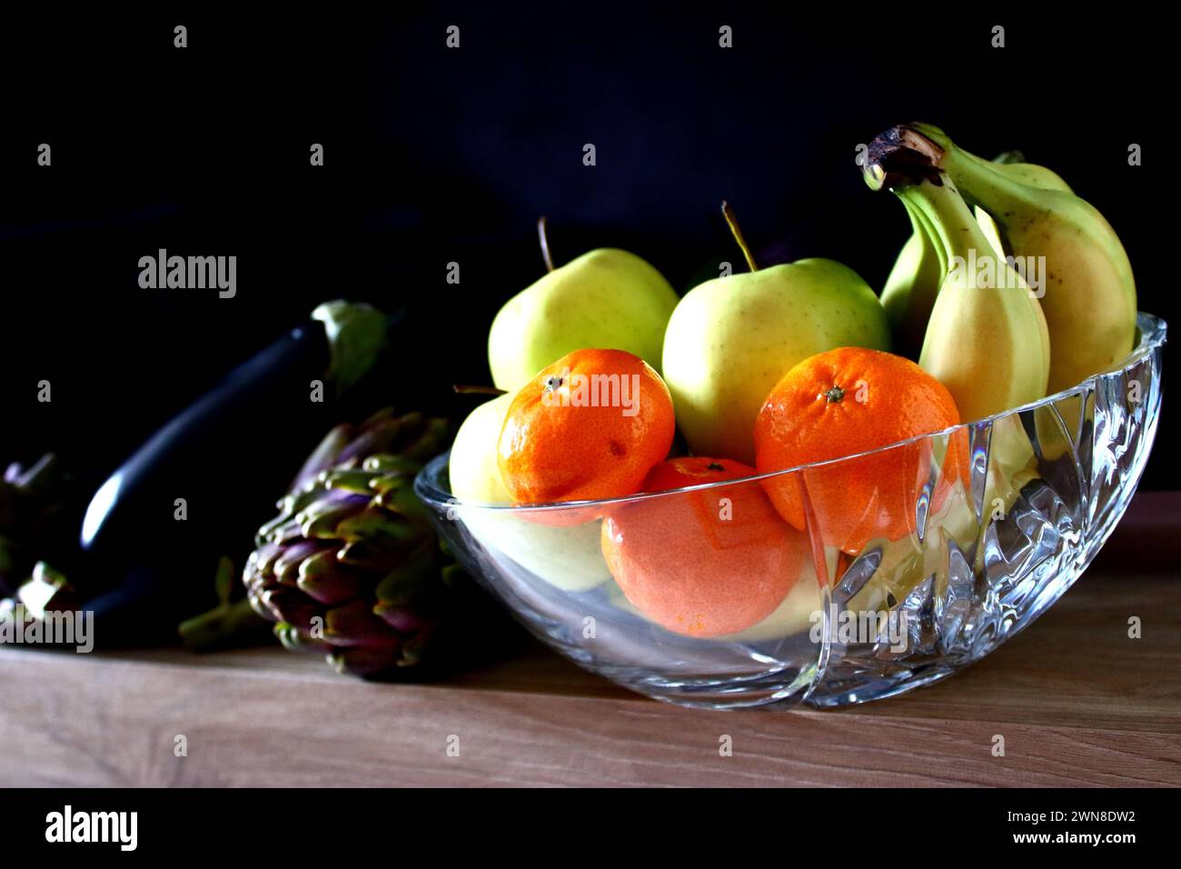 Naturaleza morta, carciofo, melanzana, mandarino, mela, banana in contenitore cristallo, fondo scuro, base foto legno, luce lato, fotografía de naturaleza muerta. Foto de stock