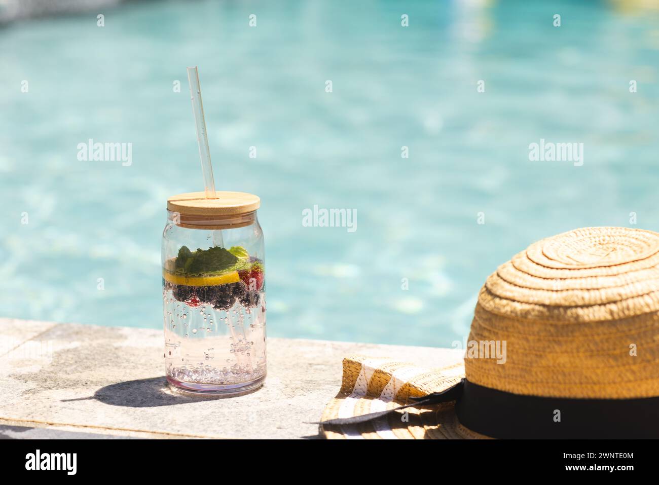 Un frasco de agua con infusión de frutas se encuentra junto a un sombrero de paja junto a una piscina Foto de stock