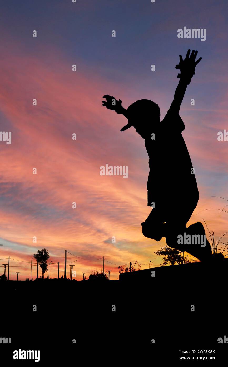 silueta de skater chico con gorro saltando en perfil en el aire con las manos en el aire con una puesta de sol en el fondo Foto de stock