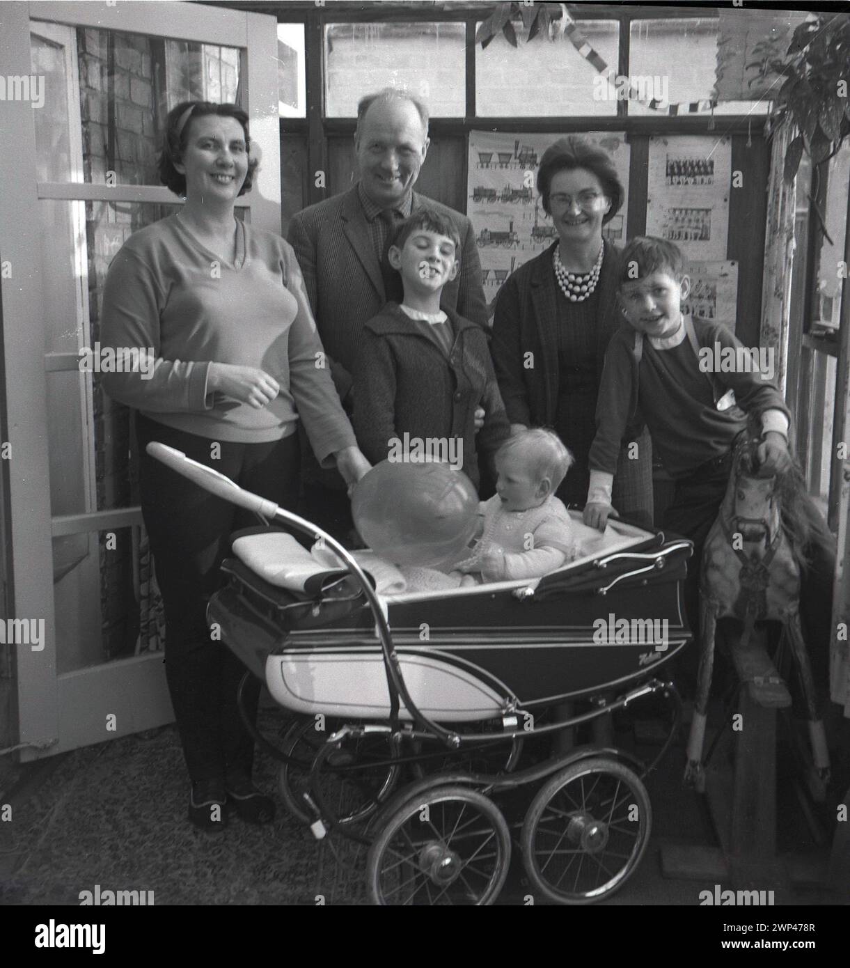 1960s, sonrisas históricas y grandes en todo momento mientras una familia se coloca para una foto dentro de una parte trasera de la casa inclinada con un niño sentado en un cochecito construido por un entrenador sosteniendo un globo, Inglaterra, Reino Unido. Cumpleaños en el techo. Foto de stock