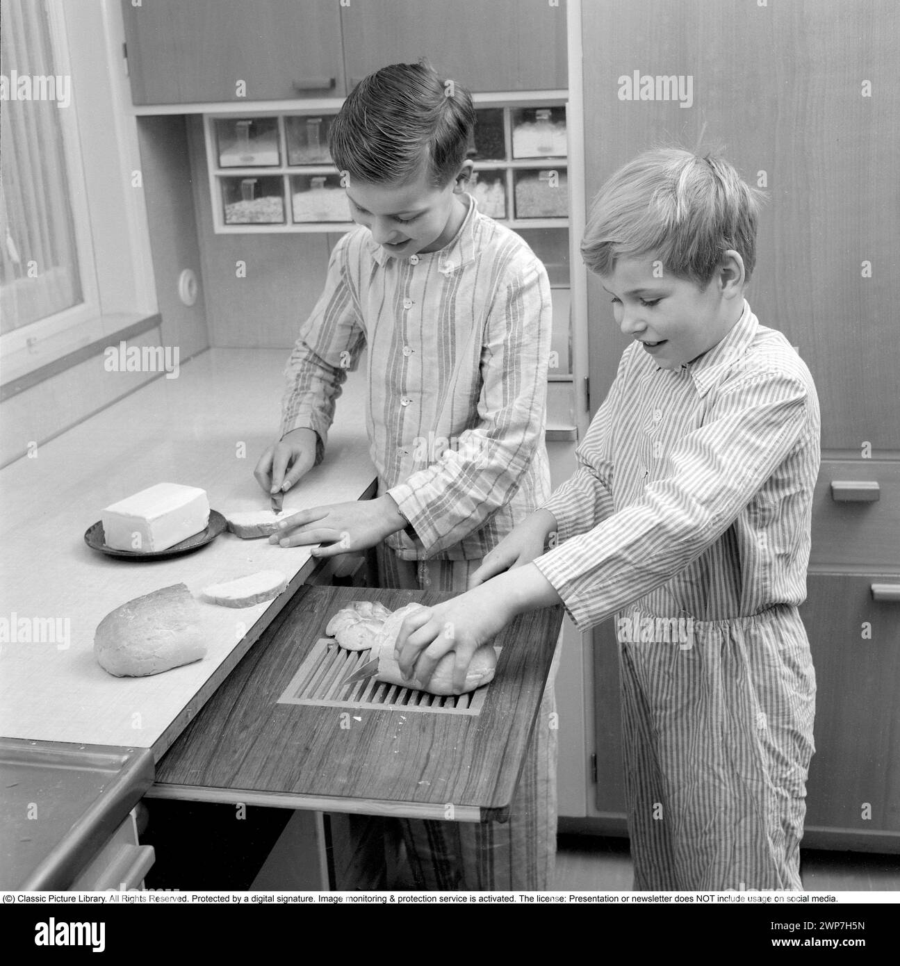 Desayuno en los años 1950 Dos niños están de pie en pijama preparando el desayuno. Uno de ellos corta el pan en la tabla de cortar y uno extiende la mantequilla en el sándwich. 1956. Conard ref 3167 Foto de stock